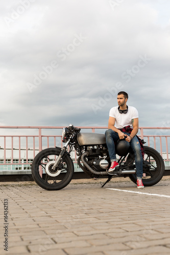 Rider guy with beard and mustache in red sneakers and white t-shirt sit on classic style biker cafe racer motorcycle. Bike custom made in vintage garage. Brutal fun urban lifestyle. Outdoor portrait.