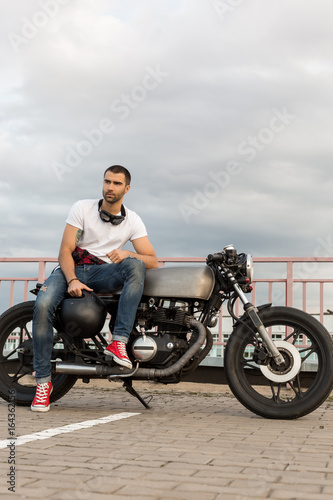 Handsome rider man with a beard and mustache sit on classic style biker cafe racer motorcycle and and wait girlfriend. Bike custom made in vintage garage. Brutal fun urban lifestyle. Outdoor portrait.