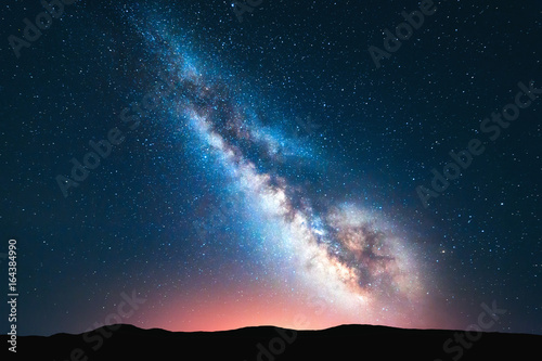 Naklejka Droga Mleczna. Fantastyczny nocny krajobraz z jasną mleczną drogą, niebem pełnym gwiazd, żółtym światłem i wzgórzami. Błyszczące gwiazdy. Malownicza scena z naszym wszechświatem. Tło przestrzeni. Niesamowita astrofotografia