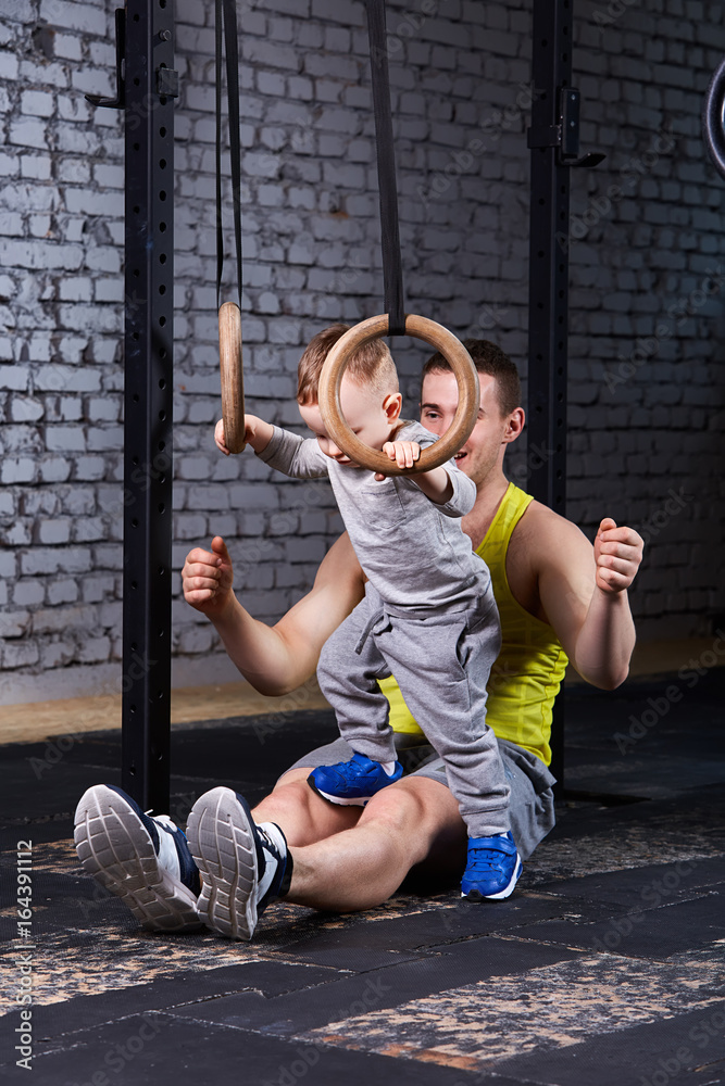 Happy father and little cute son exercising with rings against brick wall.