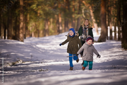Happy family walking in a winter forest. Young woman, cute toddler boy and kid boy smiling and running on the snow on a sunny day.