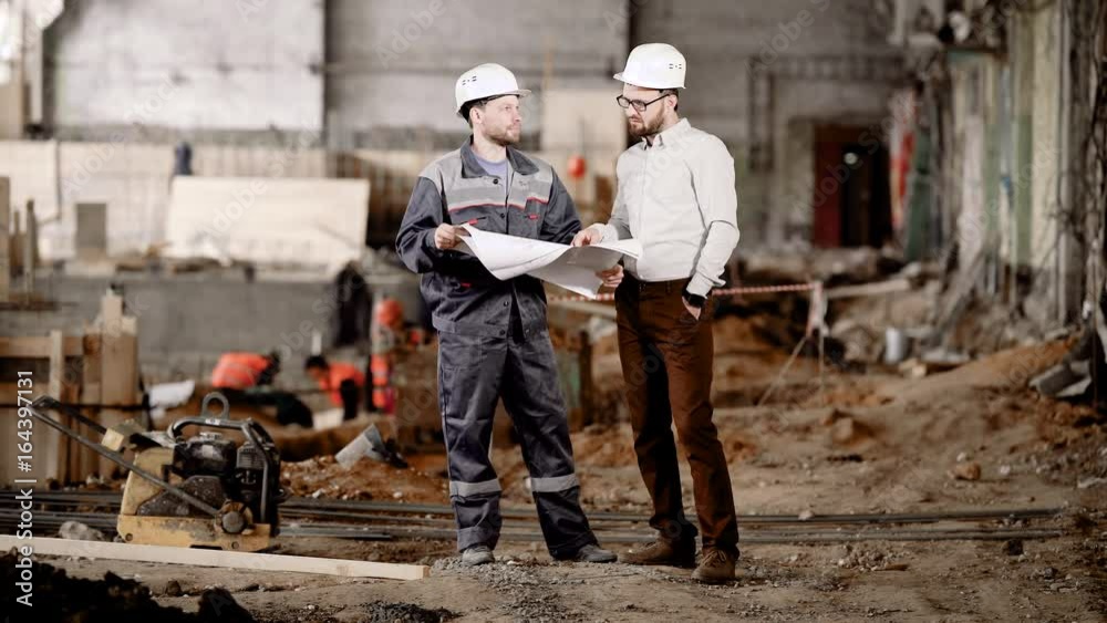 Two men in safety helmets standing in construction area and looking at scheme of future building. Foreman pointing to drawing and explaining to the worker highlights of current assembling. Two men in safety helmets standing in construction area and looking at scheme of future building. Foreman pointing to drawing and explaining to the worker highlights of current assembling.