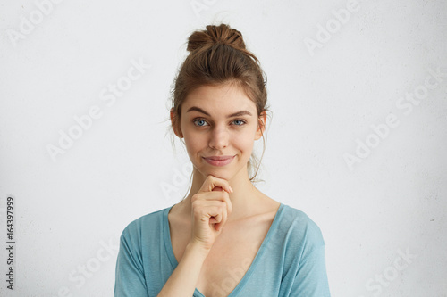 Headshot of beautiful female having cunning look raising her eyebrow and holding hand on chin having some tricky plans in her mind. Young lady looking playfully and sly isolated over white wall