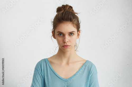 Portrait of naturally beautiful female having dark hair tied in knot wearing blue casual blouse looking seriously into camera while posing against white background. Facial expression and body language