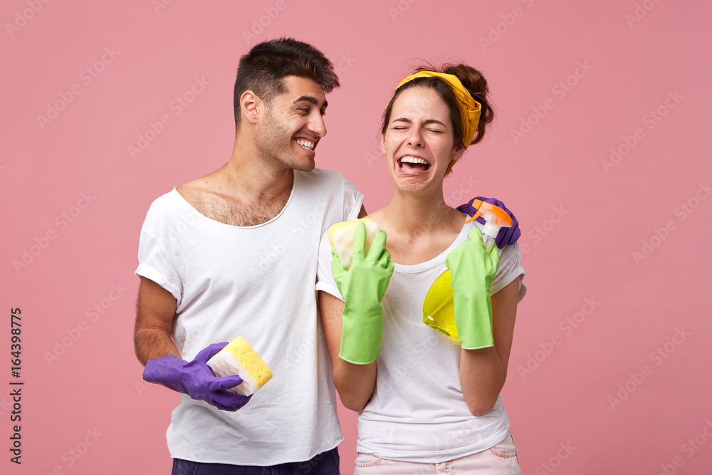 Happy young male comforting desperate stressed female in protective gloves who doesn't feel like washing dishes. Handsome positive man laughing at his sad crying girlfriend who hates cleaning up Happy young male comforting desperate stressed female in protective gloves who doesn't feel like washing dishes. Handsome positive man laughing at his sad crying girlfriend who hates cleaning up