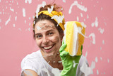 Headshot of happy beautiful positive young housewife with charming smile washing window in the kitchen, wiping out thick foam off glass surface, enjoying cleaning process, smiling at camera