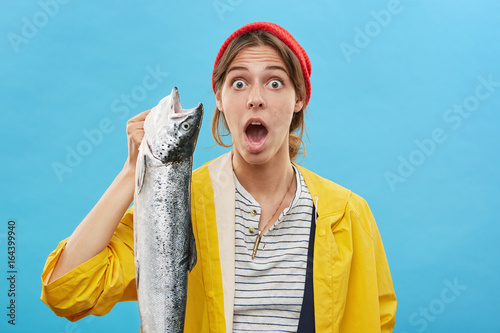 Headshot of emotional surprised young fisherwoman wearing yellow raincoat and hat holding big fish in her hand and looking at camera with mouth wide opened, shocked with fine catch. Fishing concept photo