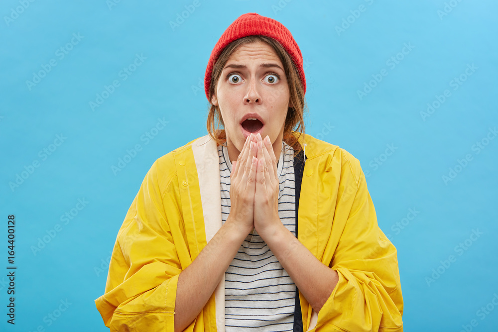 Φωτογραφία Headshot of amazed emotional young female in yellow raincoat and red hat posing against blank blue wall background with jaw dropped and eyes popped out, taken by surprise