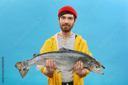 Fototapete Bearded fisherman in yellow anorak and red hat holding huge fish in hands, demonstrating his successful catch