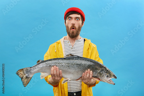 Amazement and unexpectedness concept. Shocked young fisherman with thick beard looking with bugged eyes and jaw dropped while holding huge fish not believeing that he could catch it by himself