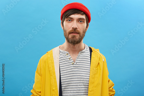 Bearded sailor dressed in red hat and yellow anorak posing against blue background. Serious man with beard having blue charming eyes dressed casually posing at studio. Fisherman going to angling photo