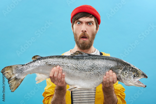 Fotografia Headshot of funny emotional young bearded Caucasian fisherman in hat and raincoa