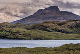 Assynt Peninsula, Scotland - June 7, 2012: Brown mountain peak under heavy brownish skies fronted by green wild rough slopes east of Loch Buine Moire. Lake waters in front.