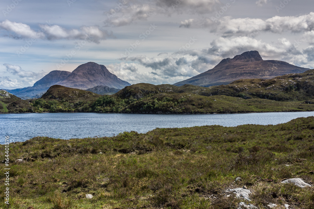 Assynt Peninsula, Scotland - June 7, 2012: Three brown mountain peaks under heavy brownish skies fronted by green wild rough slopes east of Loch Buine Moire. Lake waters in front.