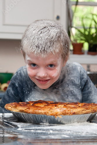 A smiling boy covered in flour and a baked pie on a table in front of him.