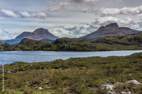 Assynt Peninsula, Scotland - June 7, 2012: Three brown mountain peaks under heavy brownish skies fronted by green wild rough slopes east of Loch Buine Moire. Lake waters in front.