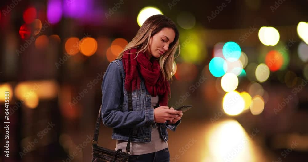 Trendy millennial girl messaging on smartphone on city street at night. Attractive Caucasian woman wearing a scarf and jean jacket, texting on cellphone downtown at night. 4k 