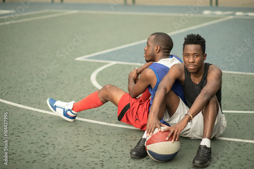 Two african american man friends sitting on street basketball court