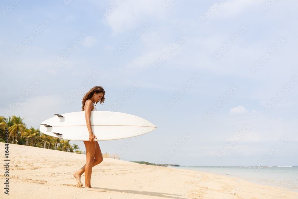 Beautiful fit surfing girl in sexy bikini swimwear with white surf shortboard surfboard blank board at sunrise or sunset on sand beach walk to ocean. Vacation concept. Summer holidays. Tourism, sport.