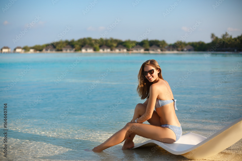 Young woman relaxing in a modern deck chair on a tropical beach with glasses on. Girl is sitting on a beach sun bed chilling near ocean, palms and bungalows. Copy space