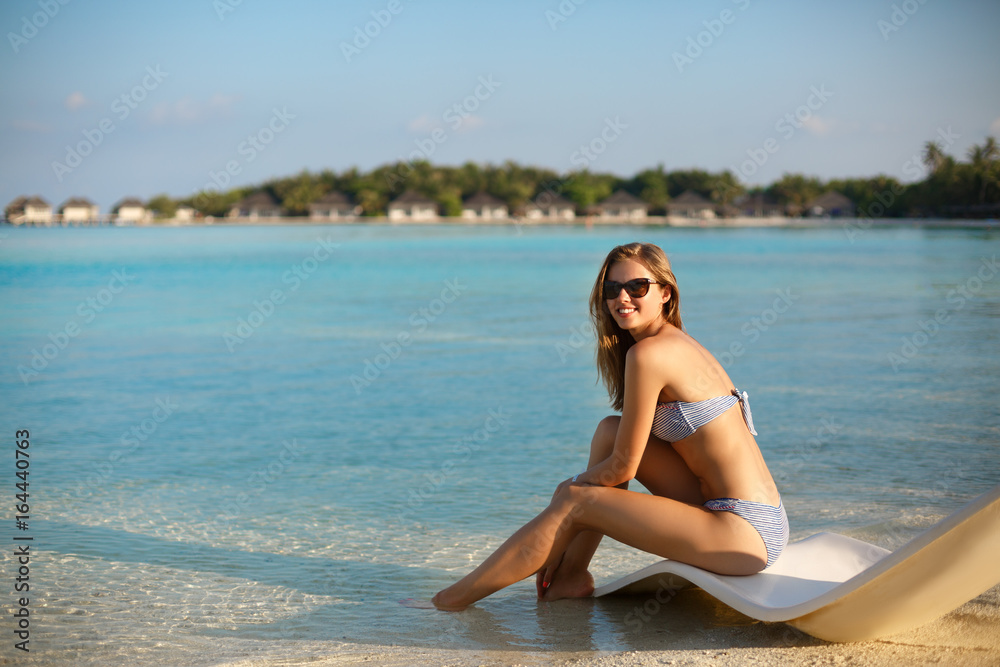 Young woman relaxing in a modern deck chair on a tropical beach with glasses on. Girl is sitting on a beach sun bed chilling near ocean, palms and bungalows. Copy space Young woman relaxing in a modern deck chair on a tropical beach with glasses on. Girl is sitting on a beach sun bed chilling near ocean, palms and bungalows. Copy space