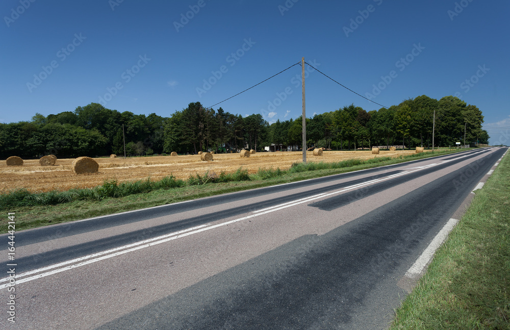 A repaired and pot hole free country road alongside a field of hay bales in Normandy, France