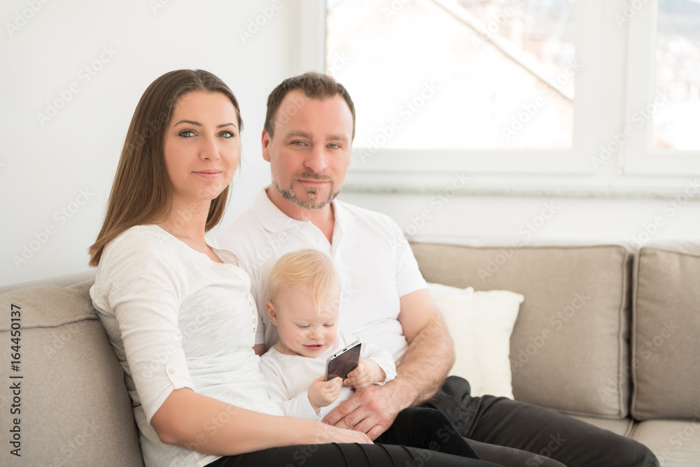 Happy family of three is enjoying at home. Parents and their beautiful baby girl sitting on the sofa and looking at the camera. Family values. Leisure together. Happy family of three is enjoying at home. Parents and their beautiful baby girl sitting on the sofa and looking at the camera. Family values. Leisure together.