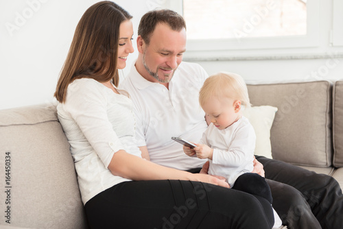 Happy family of three  is enjoying at home. Parents sitting on the sofa and looking at their beautiful baby girl playing with cell phone. Family values. Leisure together. © cirkoglu