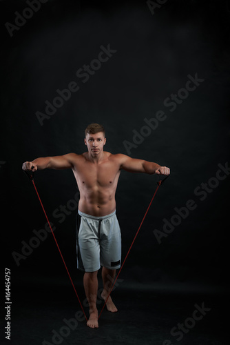 Full lenght portrait of shirtless young European bodybuilder exercising in gym, building bicep and tricep using resistance rubber band, wearing shorts. Physical training, fitness and sports concept