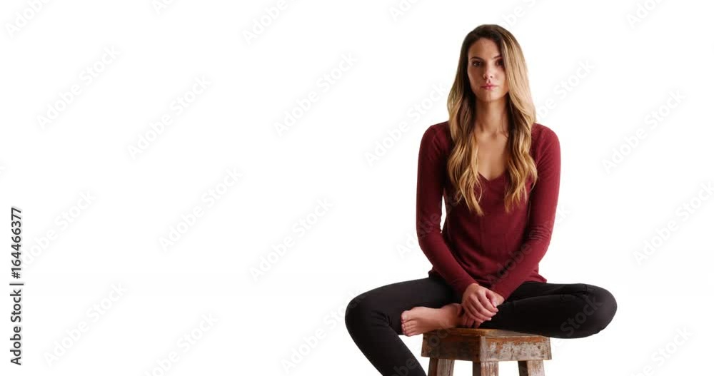 Portrait of female millennial sitting on stool, looking at camera with serious expression on white background with copyspace. Casual portrait of sad Caucasian woman sitting on chair in studio. 4k 