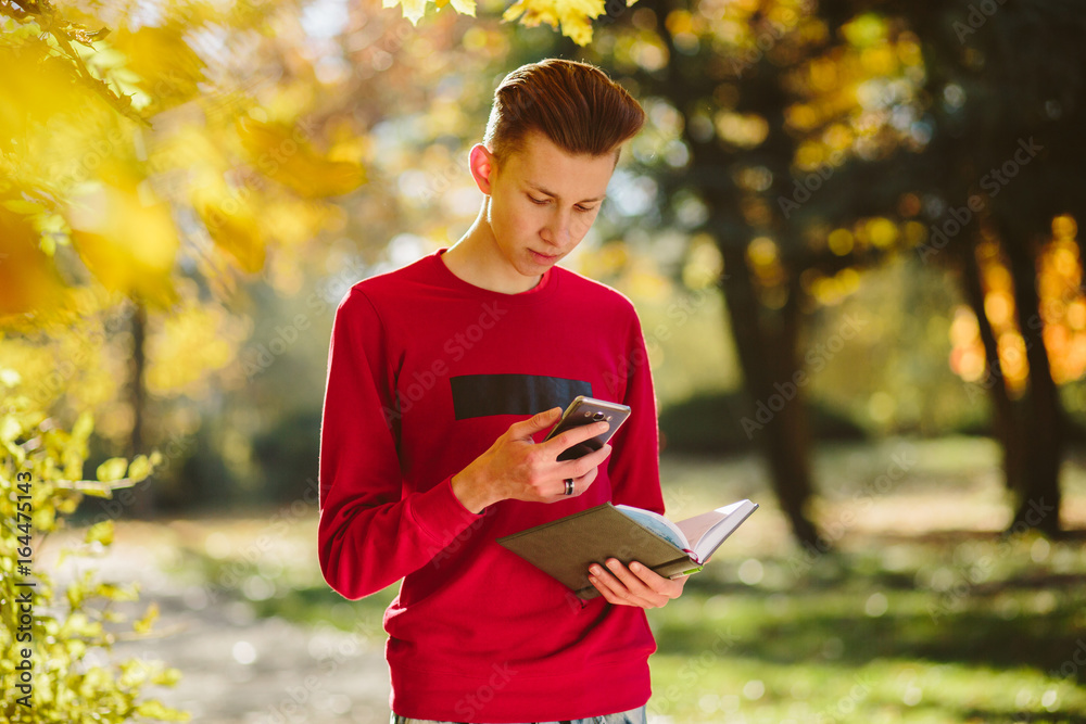 season, study, education and people concept - portrait of young hipster teenager boy in red shirt holding mobile phone and book in autumn park season, study, education and people concept - portrait of young hipster teenager boy in red shirt holding mobile phone and book in autumn park