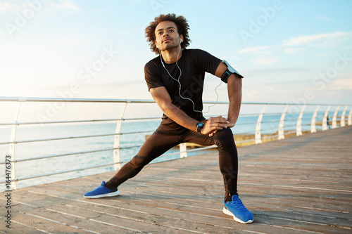 Dark-skinned male runner with beautiful fit body warming-up before cardio workout. Male athlete in sportswear stretching legs with lunge hamstring stretch exercise by the sea in morning sunlight.