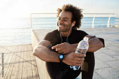 Valokuvatapetti Cheerful dark-skinned muscular athlete in black sport clothing sitting on pier after sport activities wearing white earphones