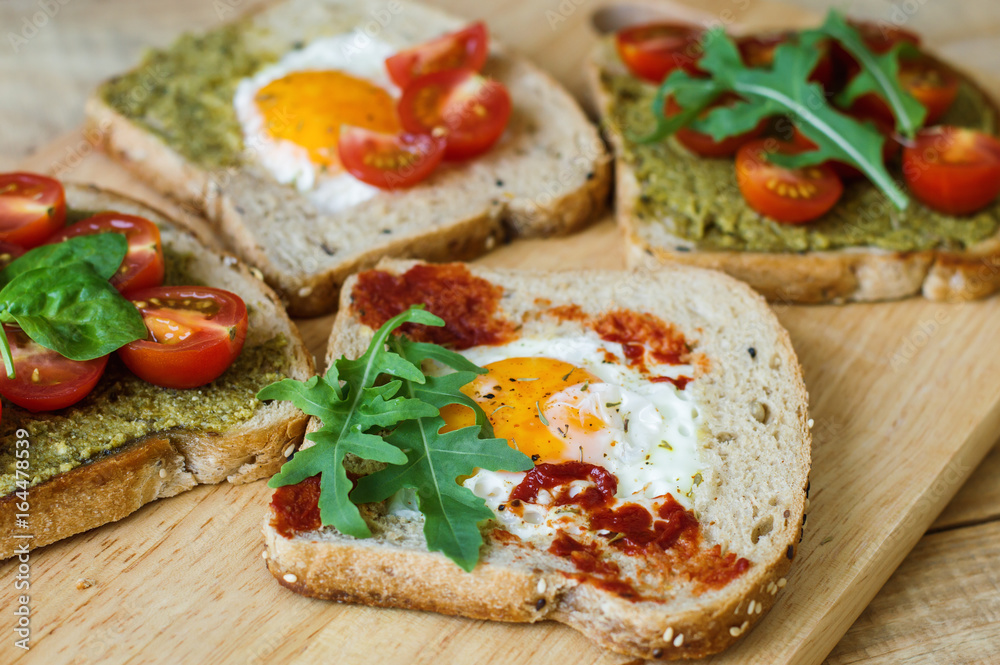 Toasts with egg, pesto and tomato sauce, arugula, spinach, cherry tomato and multi cereal bread on wooden background. Healthy breakfast. Balanced meal