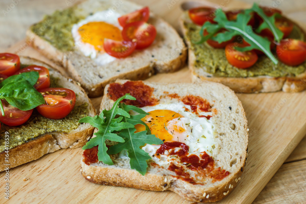 Toasts with egg, pesto and tomato sauce, arugula, spinach, cherry tomato and multi cereal bread on wooden background. Healthy breakfast. Balanced meal Toasts with egg, pesto and tomato sauce, arugula, spinach, cherry tomato and multi cereal bread on wooden background. Healthy breakfast. Balanced meal