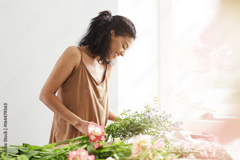 Happy tender african girl smiling making bouquet of flowers at workplace over white wall. Happy tender african girl smiling making bouquet of flowers at workplace over white wall.