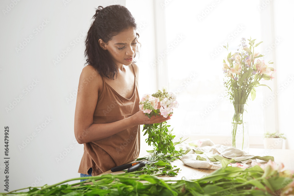 Attractive african girl florist smiling making bouquet of flowers at workplace over white wall. Attractive african girl florist smiling making bouquet of flowers at workplace over white wall.