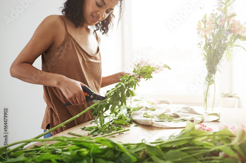 Beautiful african female florist smiling cutting stems working with flowers over white wall. photo