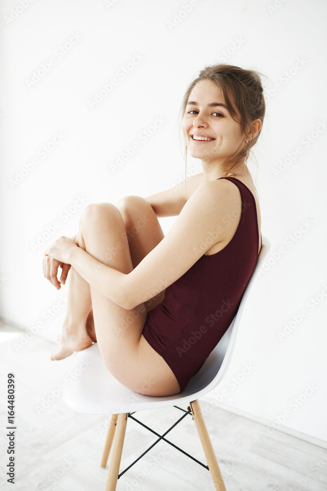 Portrait of beautiful young tender girl in swimwear smiling sitting back on chair turning to camera over white wall.