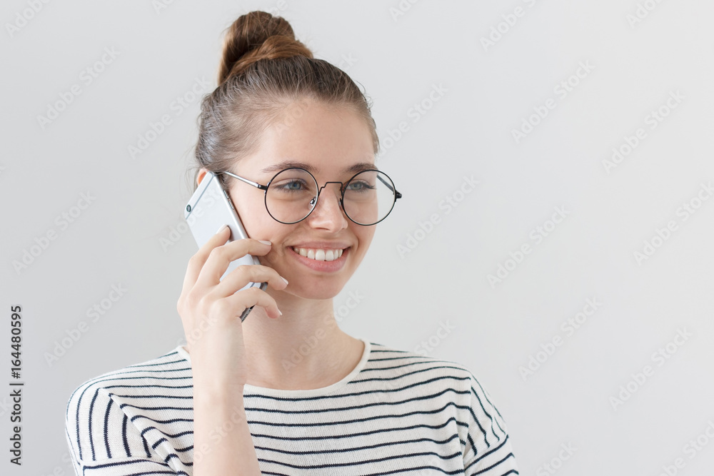 Side portrait of young European female isolated on gray background wearing hipster eyeglasses, listening to interlocutor on smartphone, showing satisfied happy smile from news she is hearing.