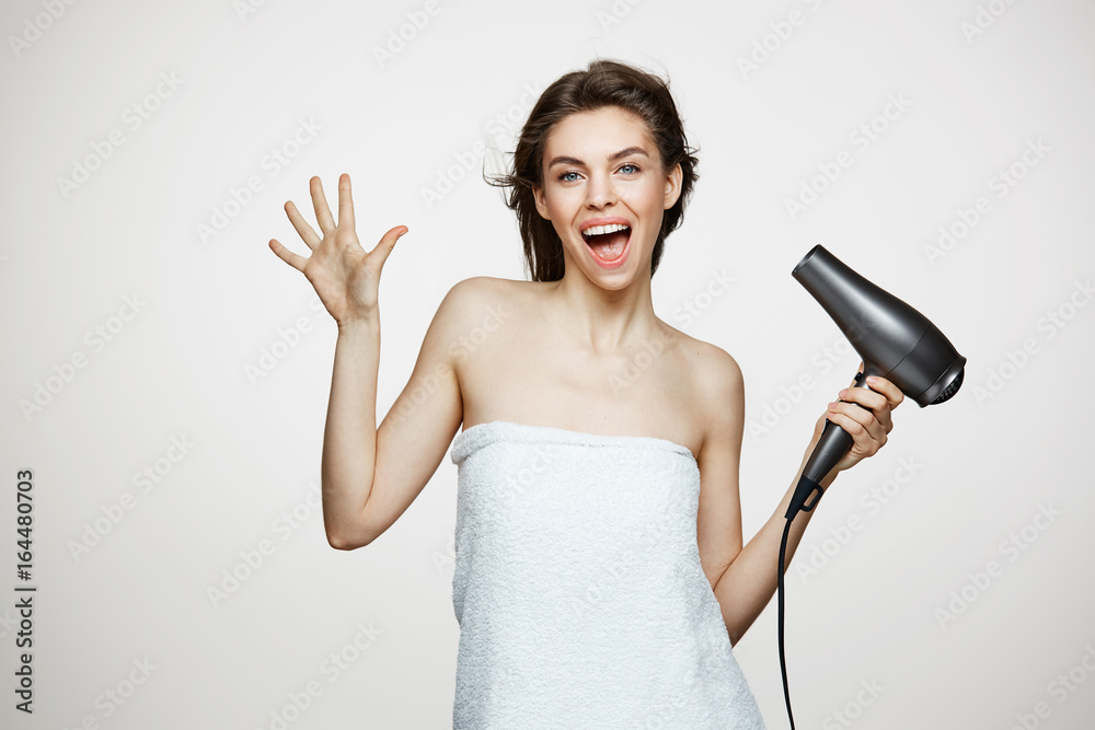 Cheerful beautiful girl in towel smiling laughing singing with hair dryer making funny face over white background. Beauty spa and cosmetology. Cheerful beautiful girl in towel smiling laughing singing with hair dryer making funny face over white background. Beauty spa and cosmetology.