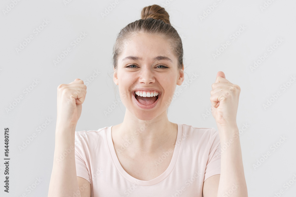 Closeup shot of positive young woman isolated on grey background sharing her triumph, raising up arms with fists, her mouth open wide while she is smiling and laughing happily and emotionally. Closeup shot of positive young woman isolated on grey background sharing her triumph, raising up arms with fists, her mouth open wide while she is smiling and laughing happily and emotionally.