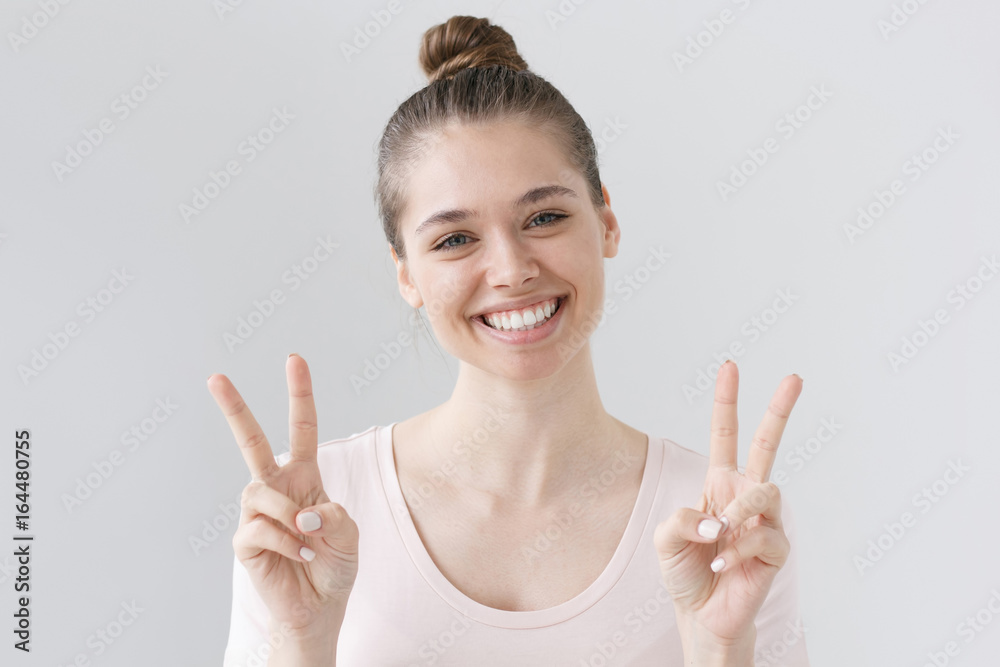 Horizontal shot of young good-looking female isolated on grey background sharing warmth and good mood by making peace gesture, laughing and smiling as if she has received heard something positive. Horizontal shot of young good-looking female isolated on grey background sharing warmth and good mood by making peace gesture, laughing and smiling as if she has received heard something positive.