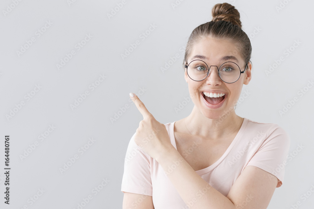 Horizontal picture of young good-looking female isolated on grey background wearing big round spectacles, pointing leftwards with forefinger, looking extremely happy, satisfied and surprised. Horizontal picture of young good-looking female isolated on grey background wearing big round spectacles, pointing leftwards with forefinger, looking extremely happy, satisfied and surprised.