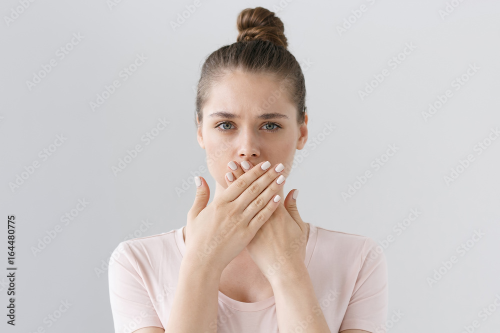 Horizontal photo of young European female isolated on gray background with expression of secrecy and mistrust as she is covering mouth with two hands not willing to disclose something important. - obrazy, fototapety, plakaty 