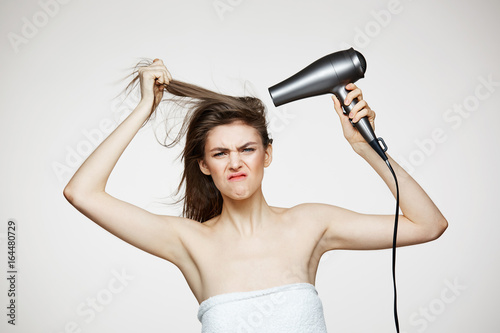 Cheerful beautiful girl in towel smiling laughing singing with hair dryer making funny face over white background. Beauty spa and cosmetology.