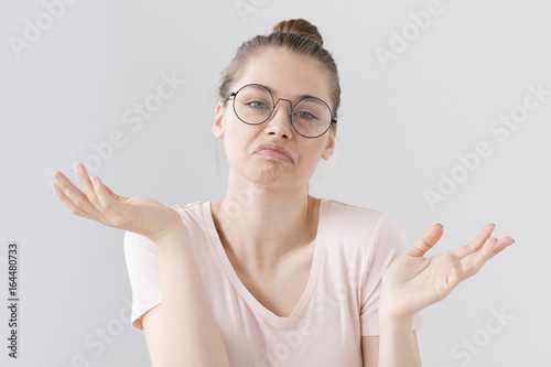Indoor photo of confused young female wearing trendy hipster eyeglasses isolated on grey background expressing helplessness and indifference with arm gesture, mouth curved in uncertain expression.