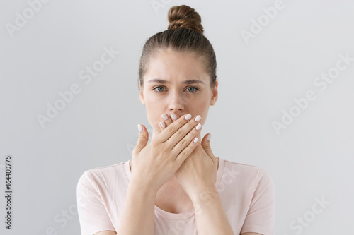 Fototapeta Horizontal photo of young European female isolated on gray background with expression of secrecy and mistrust as she is covering mouth with two hands not willing to disclose something important