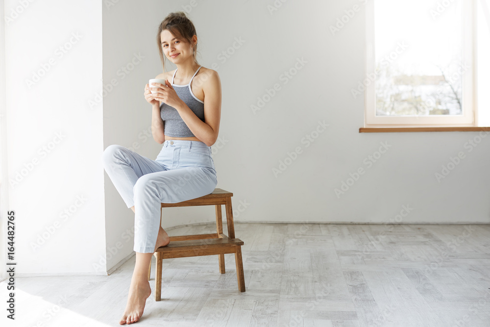 Young tender brunette girl smiling holding cup looking at camera sitting on chair over white wall early in morning.