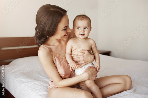 Young tender happy mother hugging her newborn baby smiling sitting on bed in morning.